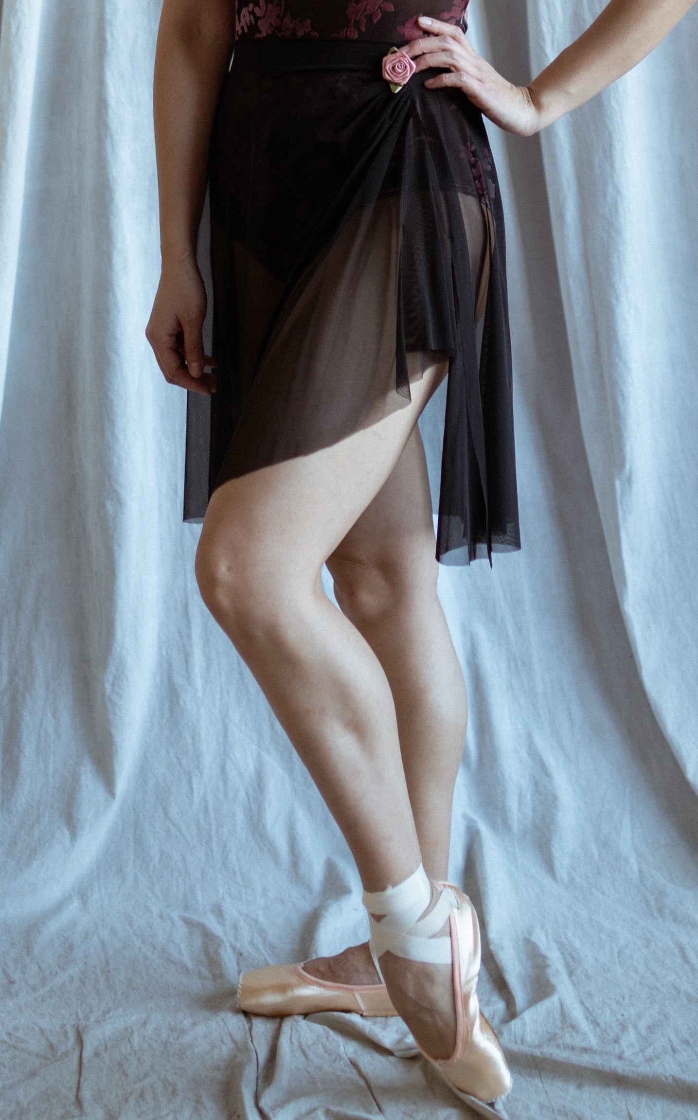 Dancer in a black mesh skirt with rose detailed posing against a draped white curtain.