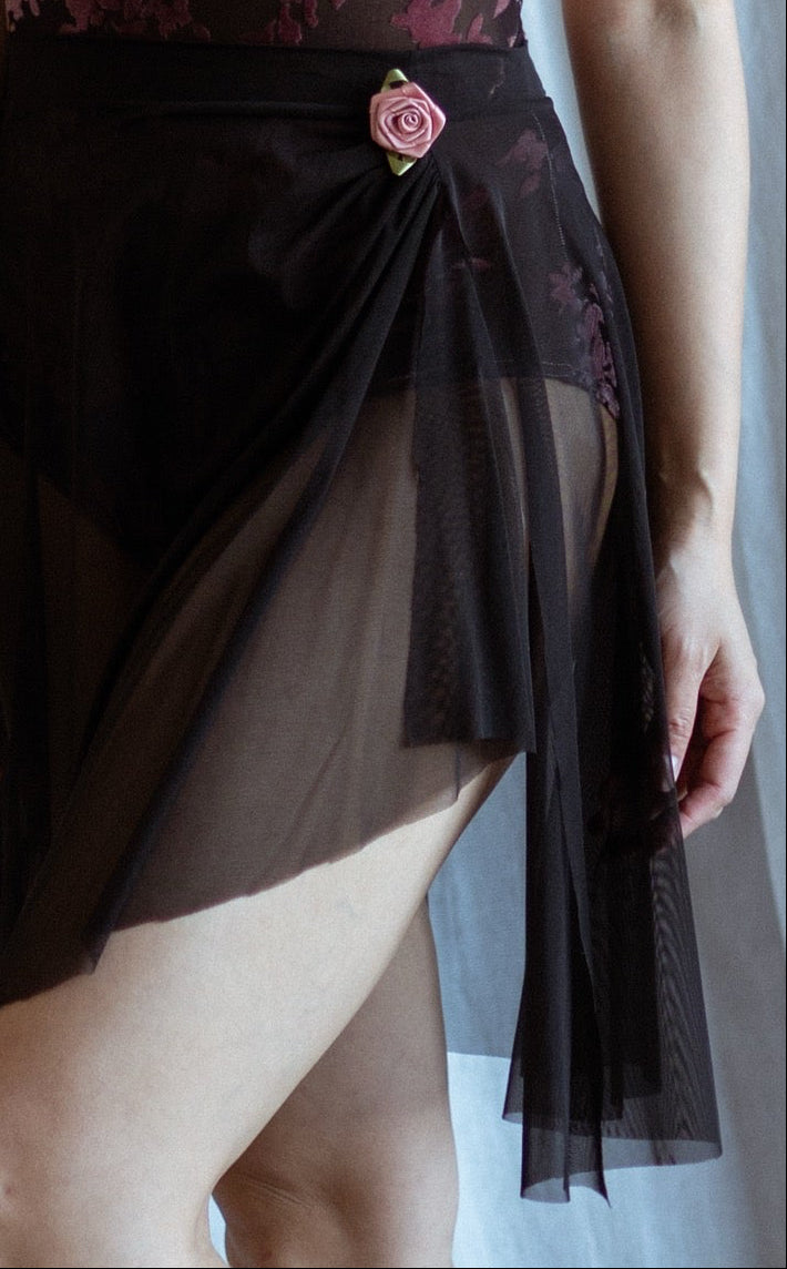 Dancer in a black mesh floral skirt standing against a draped white curtain.