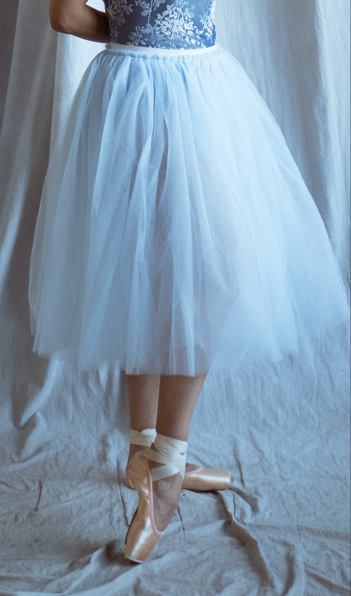 Ballerina in a blue velvet short sleeves leotard and light blue tutu skirt standing in front of draped fabric