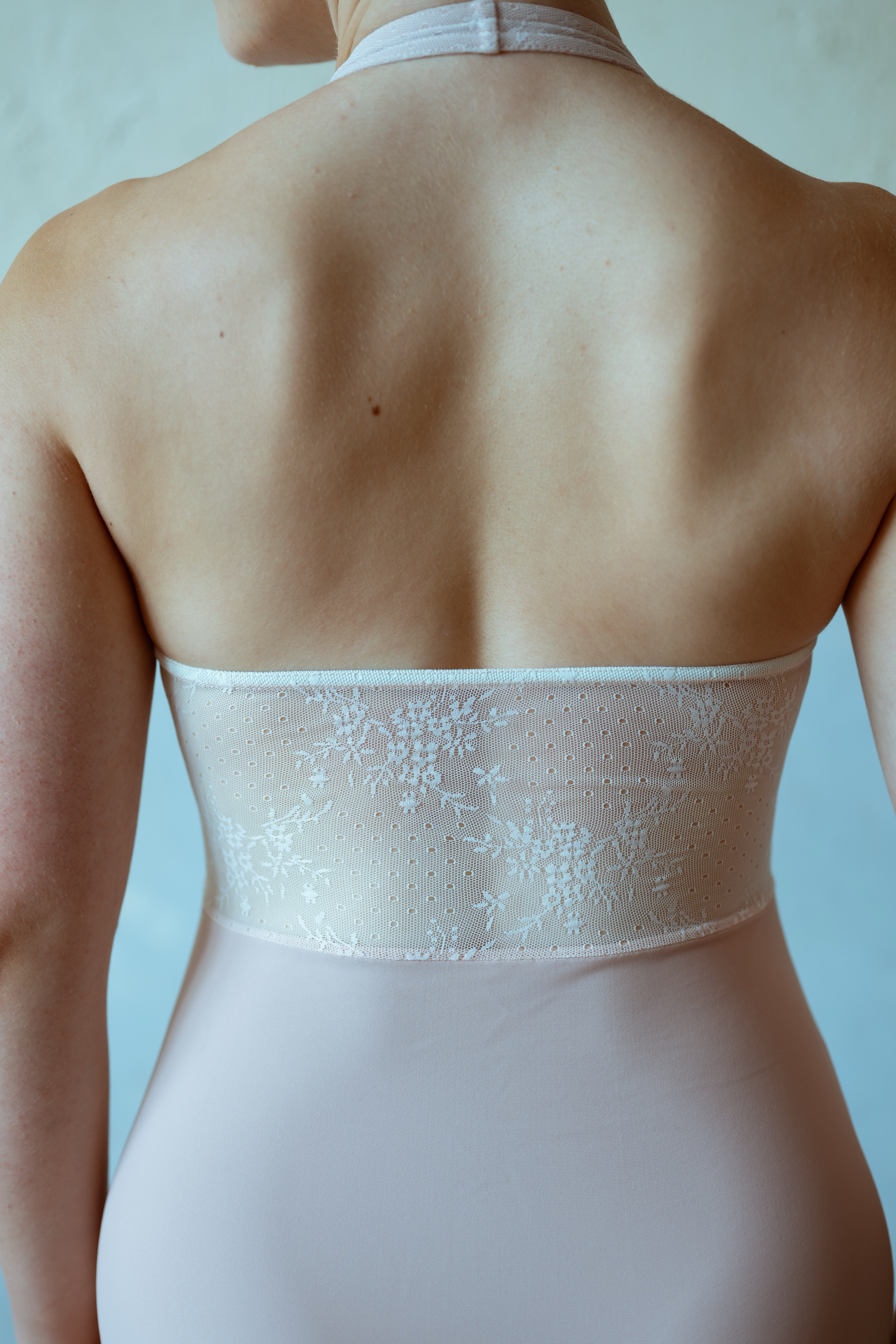 Back view of a woman wearing a dusty pink strapless bodysuit or leotard with a sheer floral lace band across the upper back and waist.