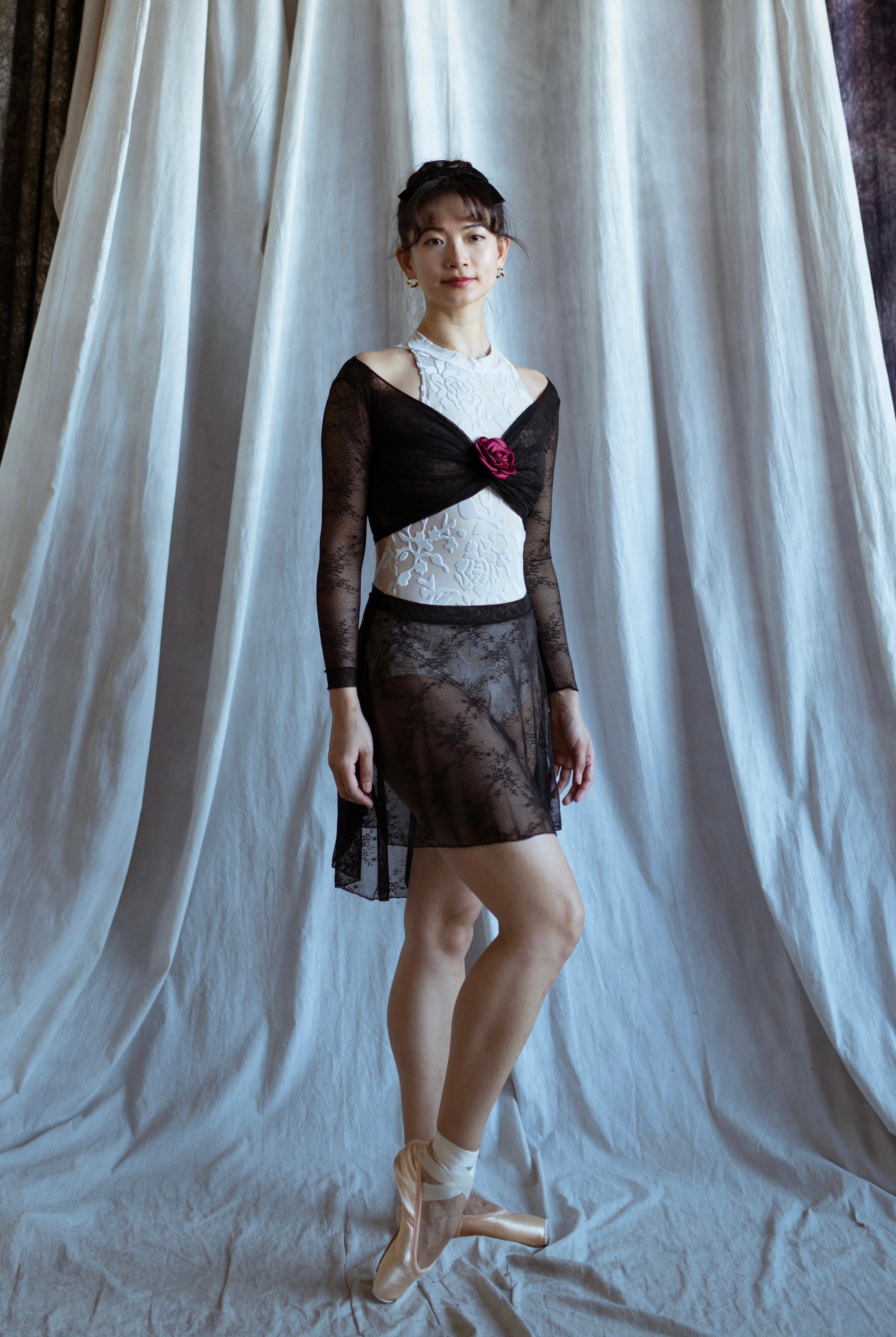 A ballerina wearing a black lace top and white leotard with a red flower and black lace skirt against a white curtain background
