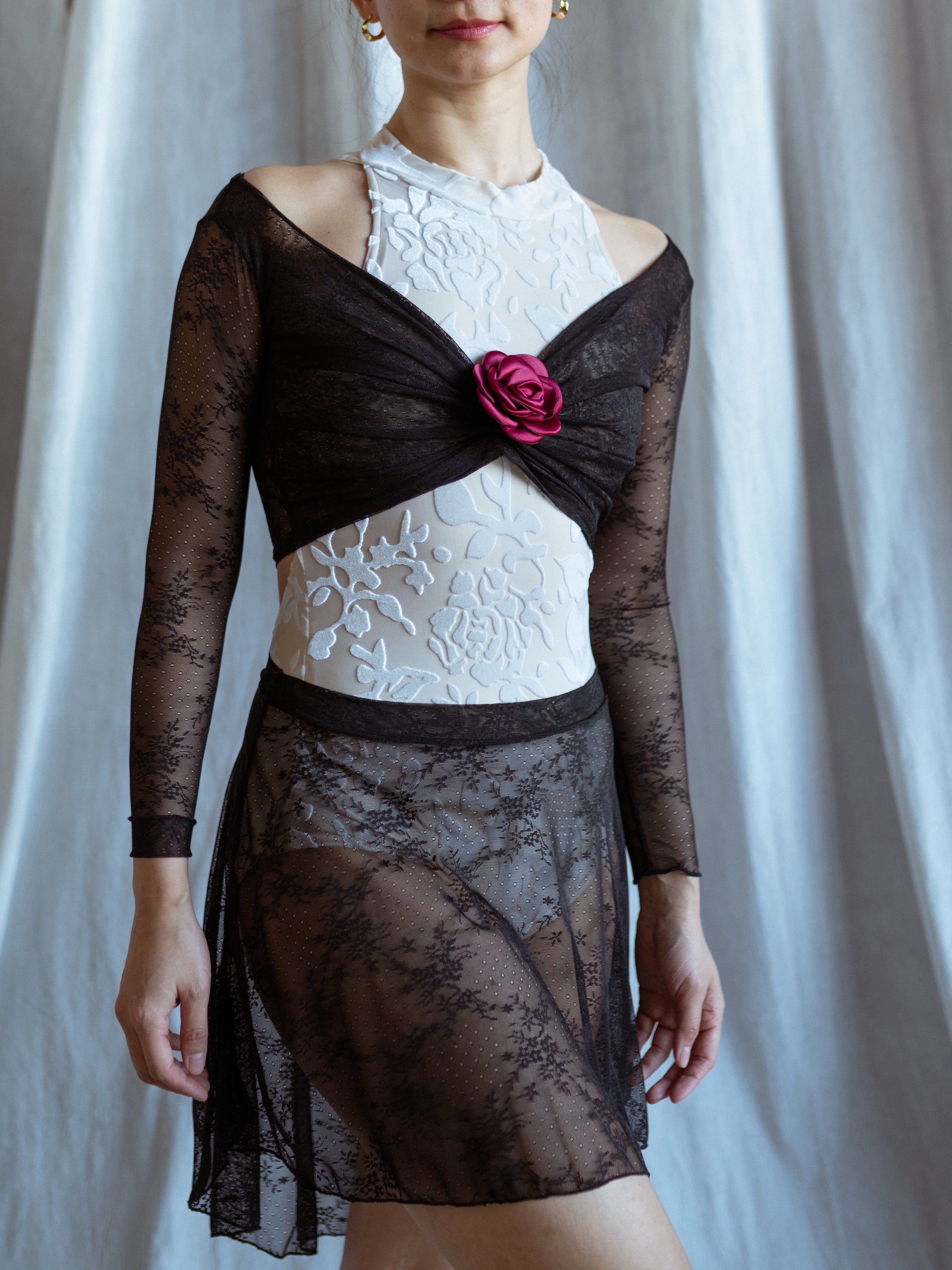 Woman wearing a black lace  top and black lace skirt with a white bodysuit against a white curtain background