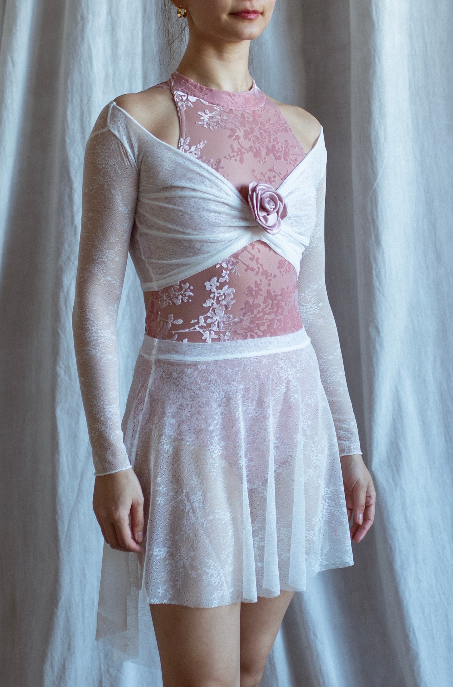 Woman wearing a pink bodysuit and white lace top with a floral detail and white lace skirt against a white curtain background