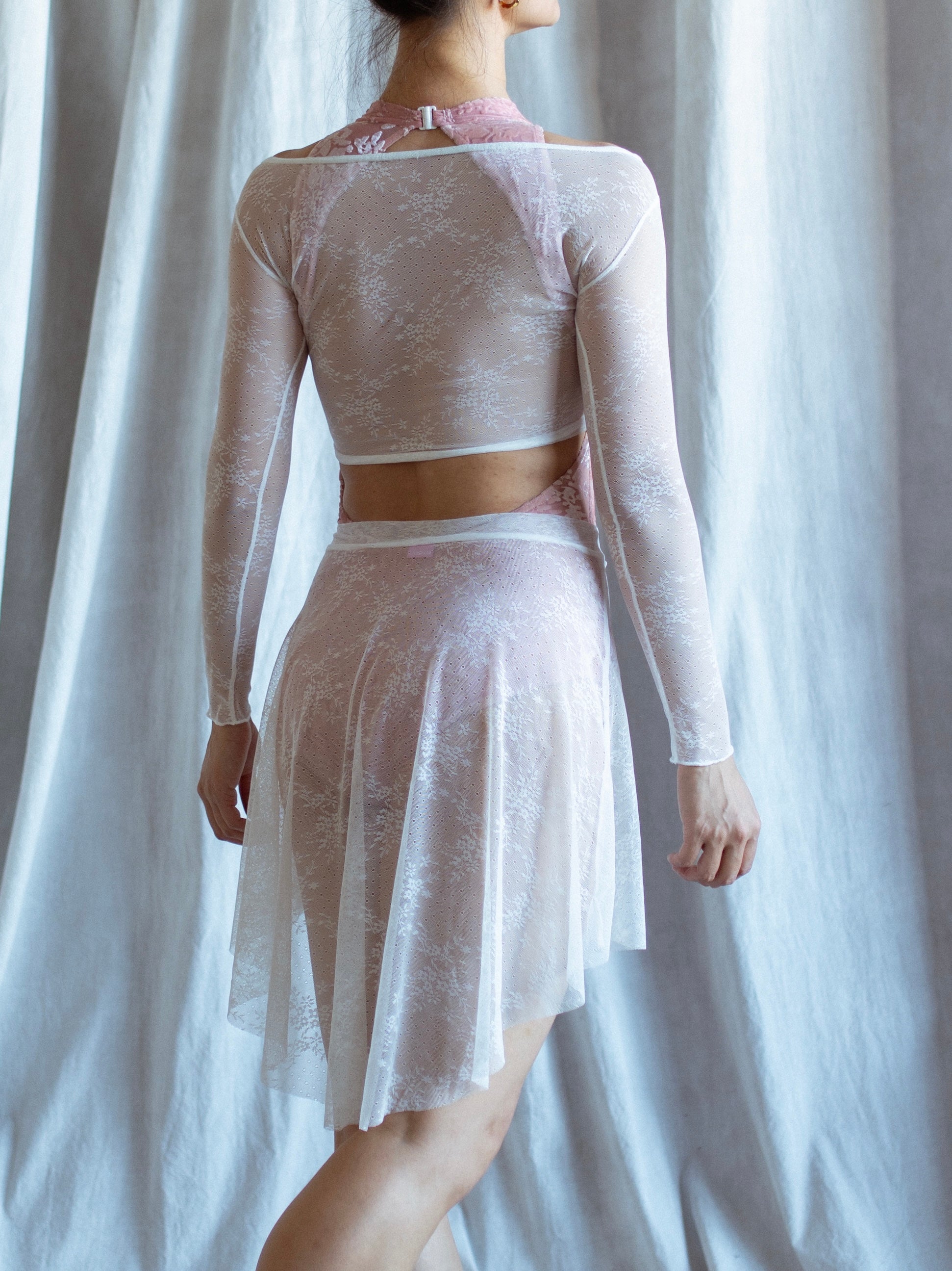 woman wearing a lace white leotard with white long sleeves lace top and a white lace skirt against a white curtain background.