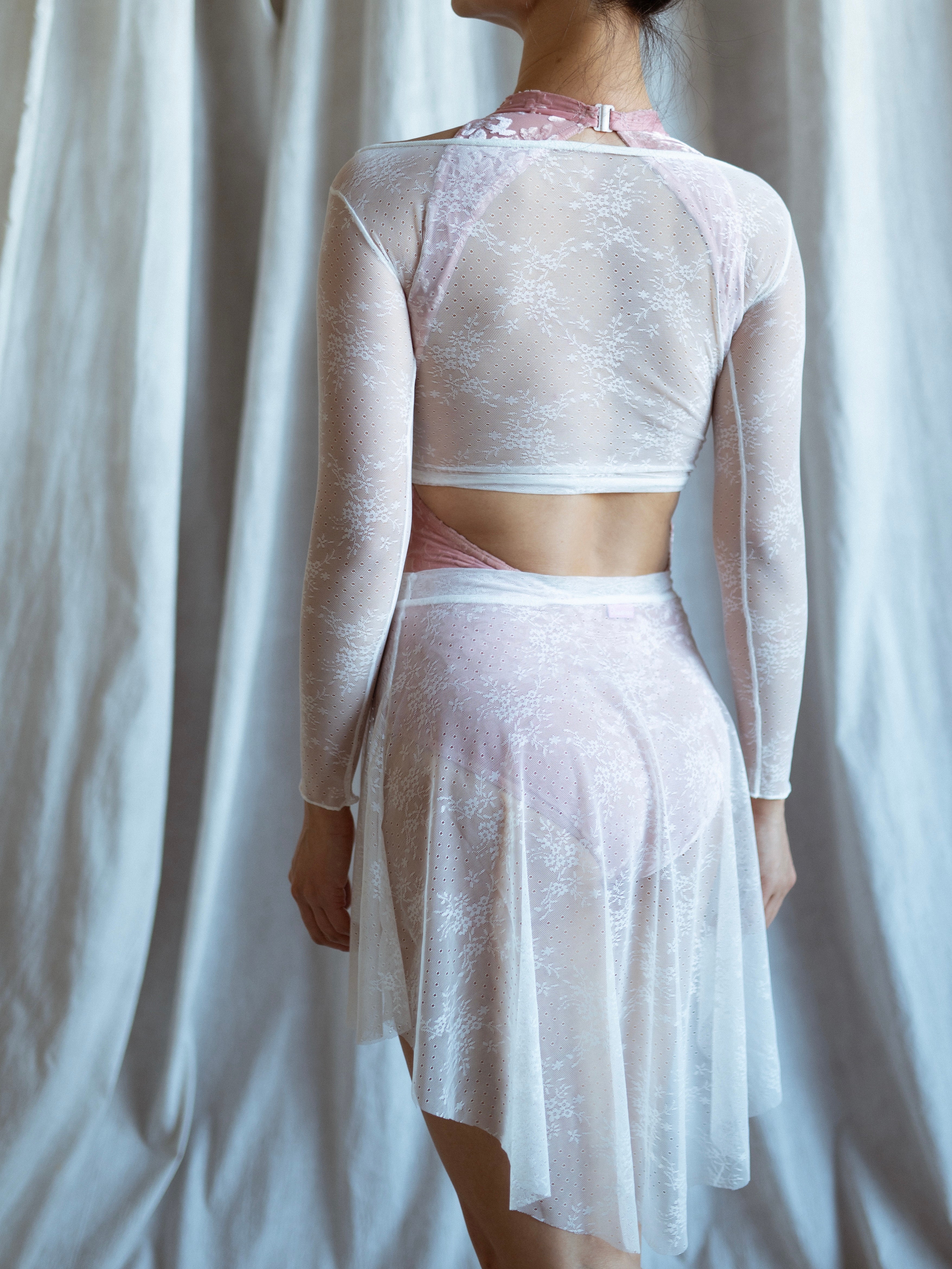 A ballerina wearing a lace white lace warm up top and pink leotard and white lace skirt against a white curtain background