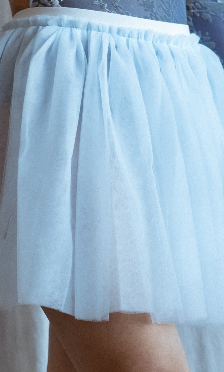 Dancer in a blue floral velvet leotard and light blue tutu skirt standing against a white curtain