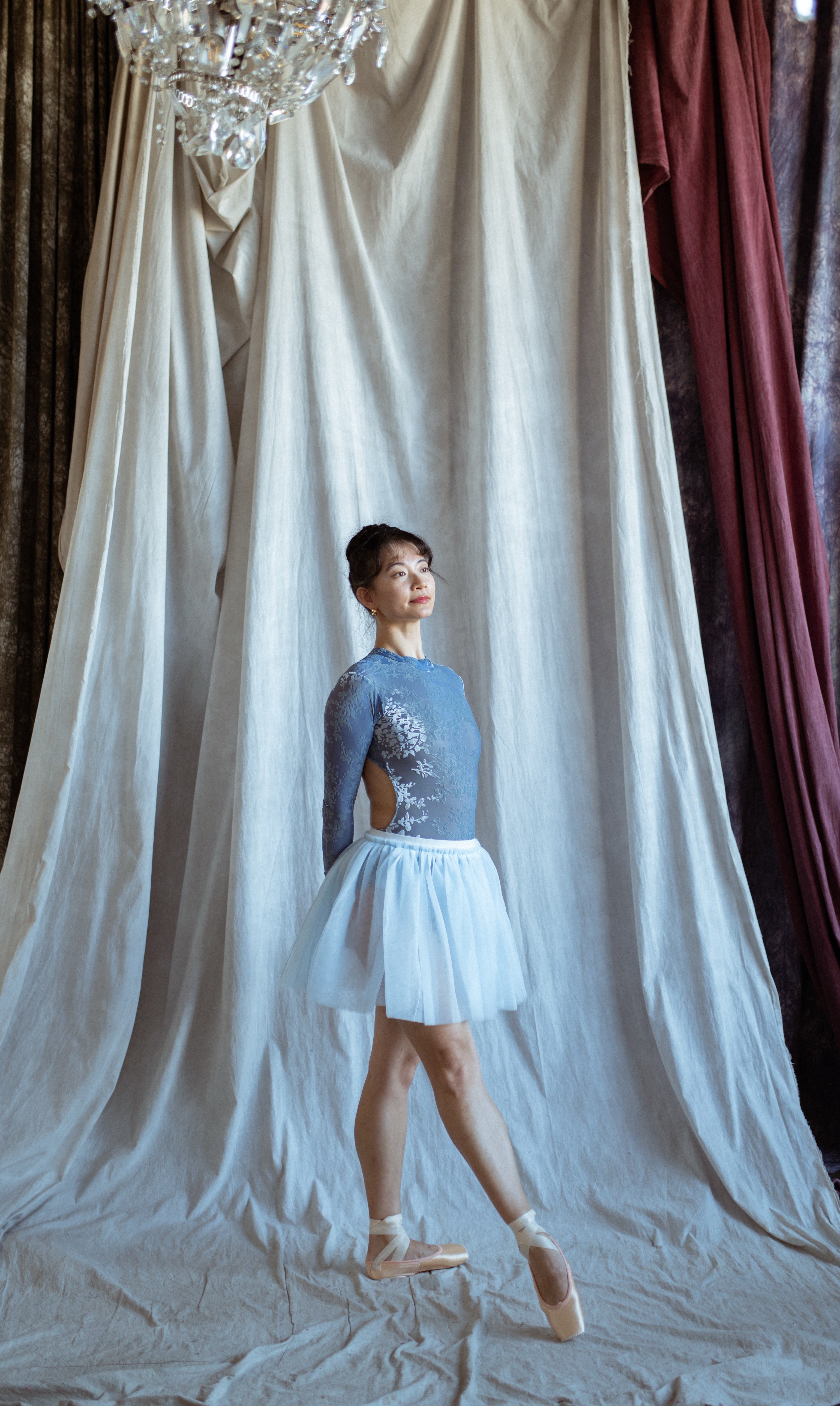 A ballerina in a blue floral velvet  long sleeves leotard and sky blue tutu  skirt standing in front of draped fabric with a chandelier above.